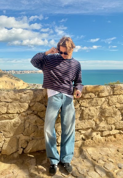 Persona ajustándose las gafas de sol, de pie junto a una pared de piedra con vista a una ciudad costera y al océano bajo un cielo azul parcialmente nublado.