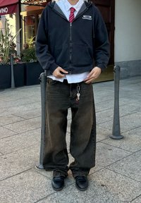 Μαύρο φούτερ με φερμουάρ πάνω από λευκό πουκάμισο και κόκκινη γραβάτα, συνδυασμένο με φαρδιά σκούρα τζιν και μαύρα παπούτσια; κλειδιά προσαρτημένα στον βρόχο της ζώνης.