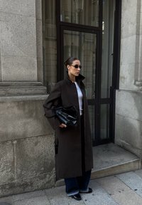 Lange bruine jas met een hoge kraag, witte top, donkere jeans en een zwarte handtas. Accessoires zijn kleine zonnebrillen en een ring.