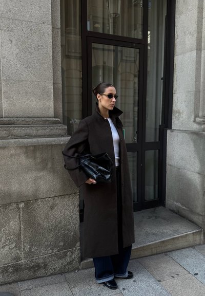 Abrigo largo marrón con un cuello alto, camiseta blanca, jeans oscuros y bolso negro. Los accesorios incluyen gafas de sol pequeñas y un anillo.