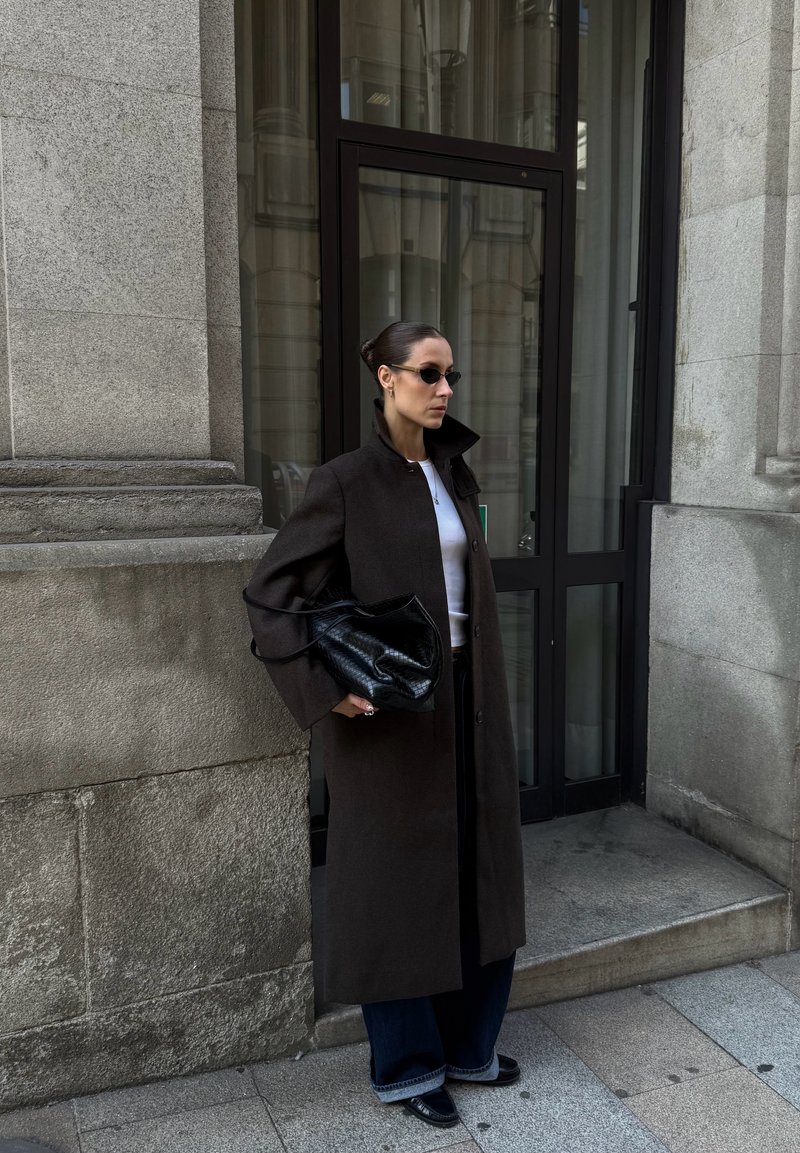 Lange bruine jas met een hoge kraag, witte top, donkere jeans en een zwarte handtas. Accessoires zijn kleine zonnebrillen en een ring.