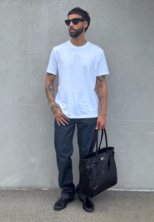 Homme avec barbe et lunettes de soleil portant un t-shirt blanc et un jean foncé, tenant un grand sac en cuir noir, debout contre un mur gris.