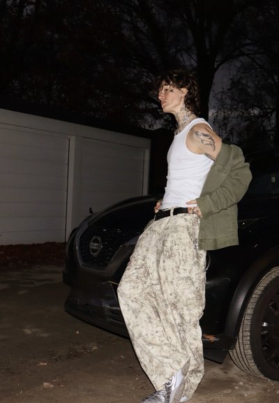 Camiseta sin mangas blanca, chaqueta oversize verde oliva y pantalones cargo claros con estampado. Zapatillas plateadas y tatuajes prominentes visibles. De fondo, un coche oscuro.