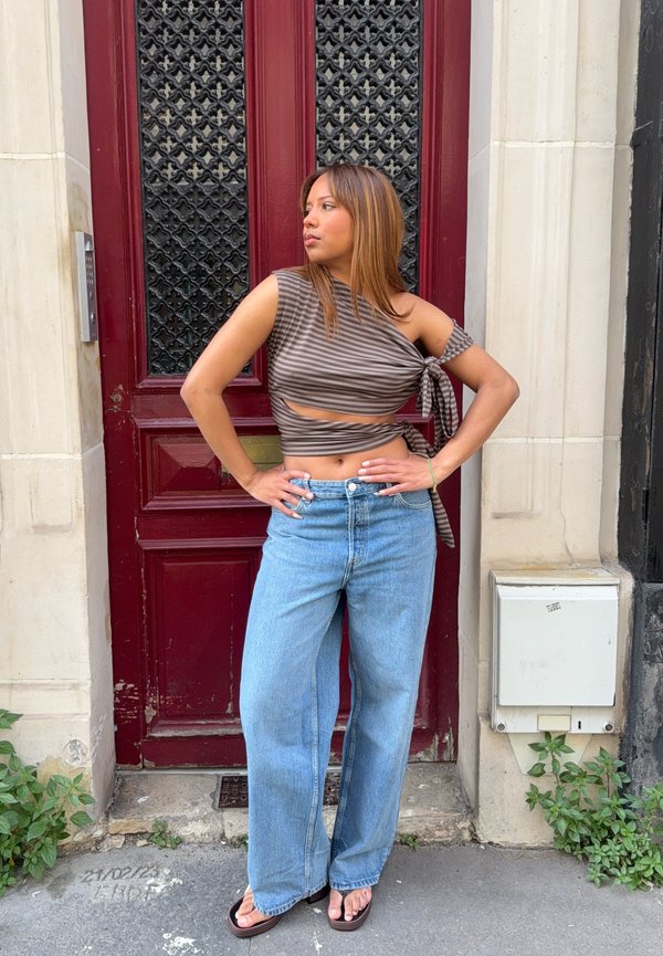 Frau in gestreiftem, einseitig schulterfreiem Crop-Top und weitem Jeanshose steht mit den Händen in der Hüfte vor einer roten Tür auf einer Stadtstraße.