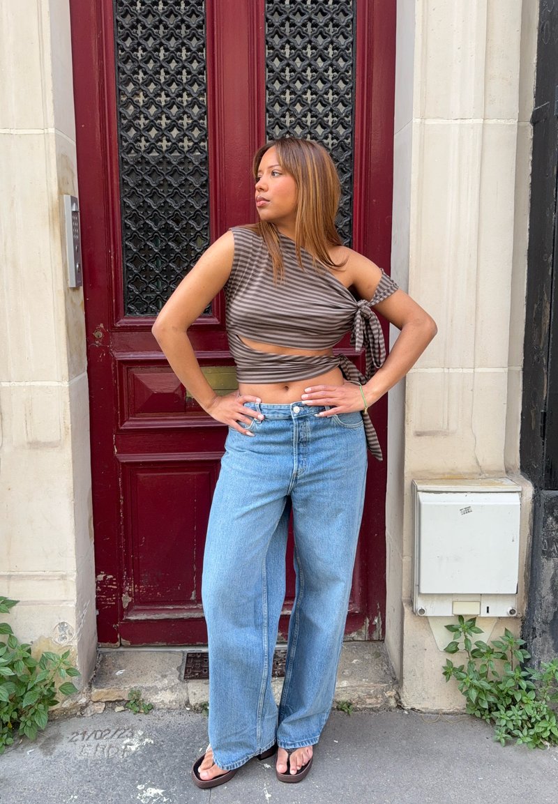 Femme en crop top rayé à une épaule et jean à jambes larges, debout les mains sur les hanches devant une porte rouge dans une rue de la ville.