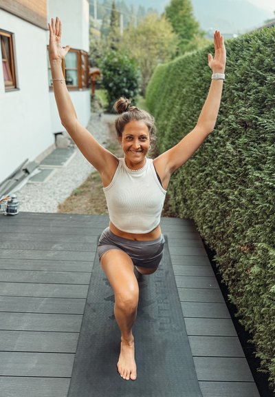 Mujer con una camiseta sin mangas blanca y pantalones cortos grises realiza una zancada sobre una esterilla de yoga negra. El fondo presenta vegetación y un edificio de madera.