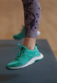 Türkise Sportschuhe mit strukturiertem Obermaterial, weißer Sohle und Schnürsenkeln. Getragen mit gemusterten lila Leggings, die ein Farbspritzdesign aufweisen.