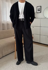 Person trägt schwarzen Cardigan, weißes T-Shirt, schwarze Hose und schwarze Schuhe und steht in einem minimalistischen Wohnzimmer mit beigem Sofa und weißem Schrank.