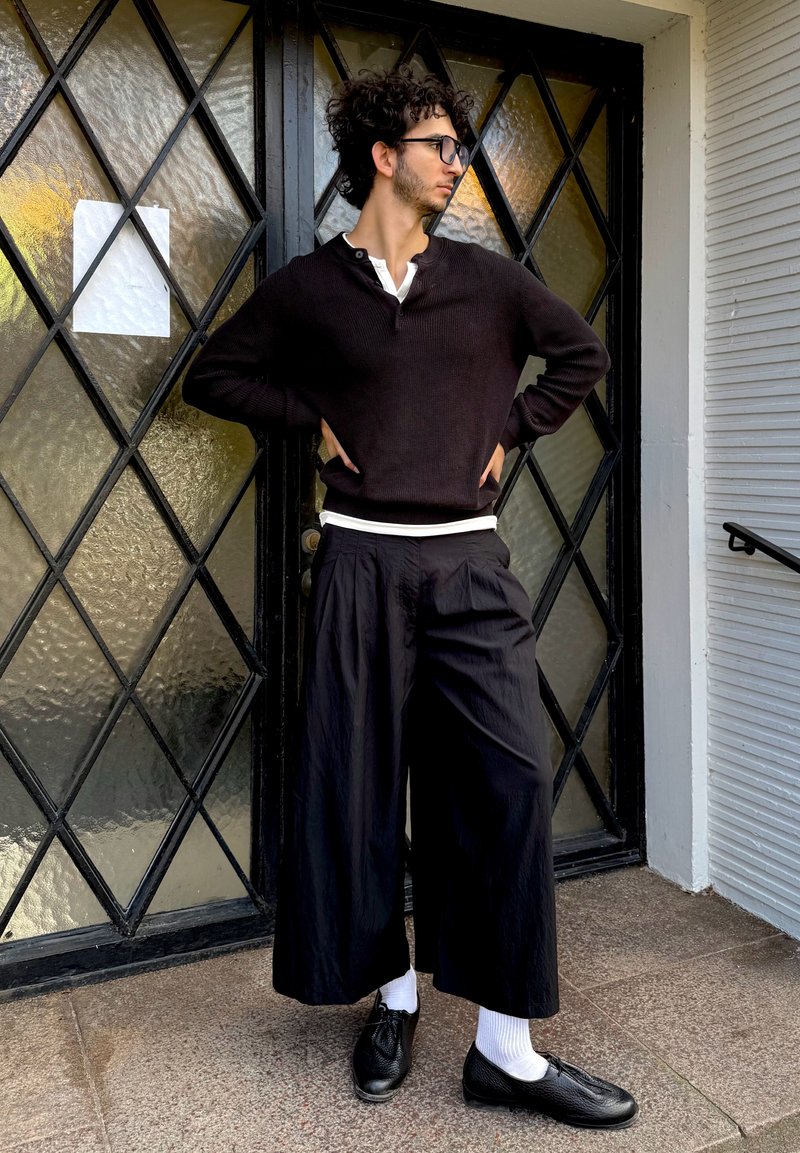 Suéter de malha preta sobre uma camisa branca, calças largas pretas, sapatos pretos e meias brancas. De pé ao lado de uma porta de vidro com padrão de diamante.