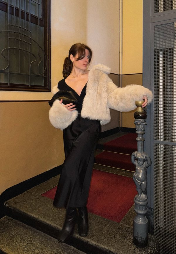 Vrouw in zwarte jurk en beige bontjas staat op trappen, houdt een zwarte clutch vast en rust haar hand op een sierlijke metalen paal.