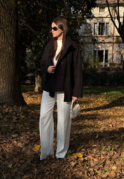 Abrigo negro extragrande, top blanco ajustado, pantalones blancos de pierna ancha y bolso blanco pequeño. De pie sobre hojas caídas con un fondo de árboles.