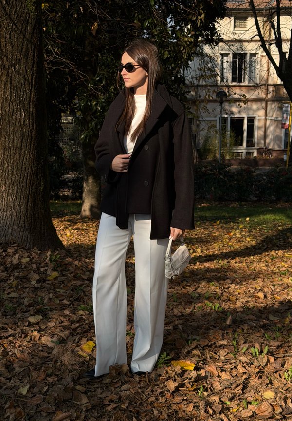 Schwarzer übergroßer Mantel, weißes figurbetontes Oberteil, weiße Hosen mit weitem Bein und kleine weiße Handtasche. Steht auf fallenden Blättern vor einem Baum.