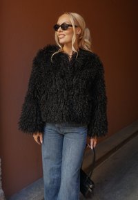 Giacca corta in pelliccia sintetica nera con maniche lunghe, finitura testurizzata. Abbinata a jeans a gamba larga azzurri e accessori neri.