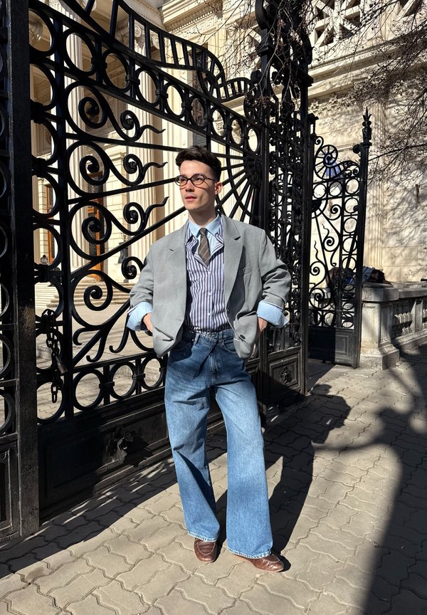 Fiatal férfi szemüvegben, szürke blézerben és csíkos ingben áll díszes fekete kovácsoltvas kapuk előtt egy napos napon.