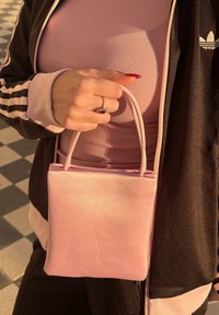 Main tenant un sac à main rose pâle par ses anses, portant une veste Adidas noire et rose ainsi qu'un haut côtelé rose, bague visible au doigt.