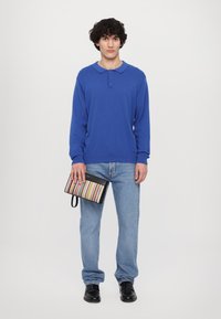 Blauwe lange mouwen polo met een kraag, gecombineerd met lichtblauwe jeans en zwarte schoenen. Een gestreepte clutch in de linkerhand.