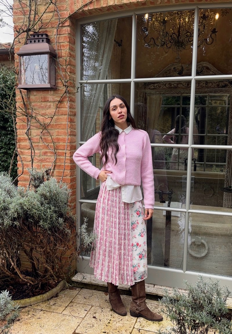 Femme aux longs cheveux foncés portant un cardigan rose, une jupe fleurie et des bottes marron, debout près d'un mur en briques et d'une porte en verre avec un lustre à l'intérieur.