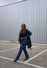Top curto preto, casaco longo preto, jeans azul folgados e botas pretas de salto alto, diante de uma parede cinza corrugada numa área de estacionamento.