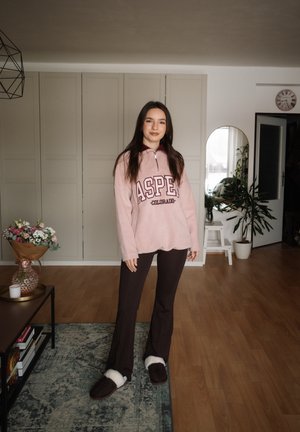 Joven mujer está de pie en la sala usando una sudadera rosa de Aspen Colorado, pantalones oscuros y pantuflas, con flores sobre la mesa y plantas cercanas.