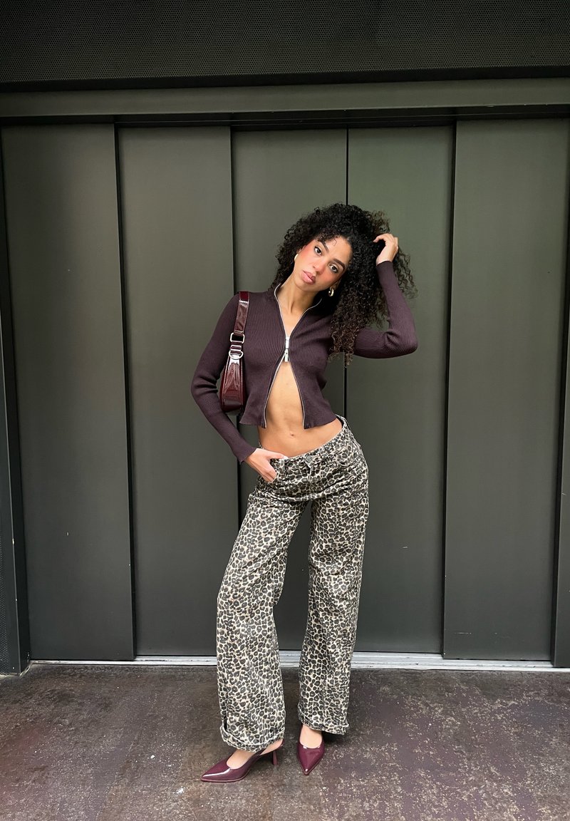 Nertas trumpas tamsiai violetinės spalvos kardiganas su užtrauktuku; plačios kelnės su leopardžio raštu; bordo aukštakulniai; pečių krepšys; tekstūruoti garbanoti plaukai.