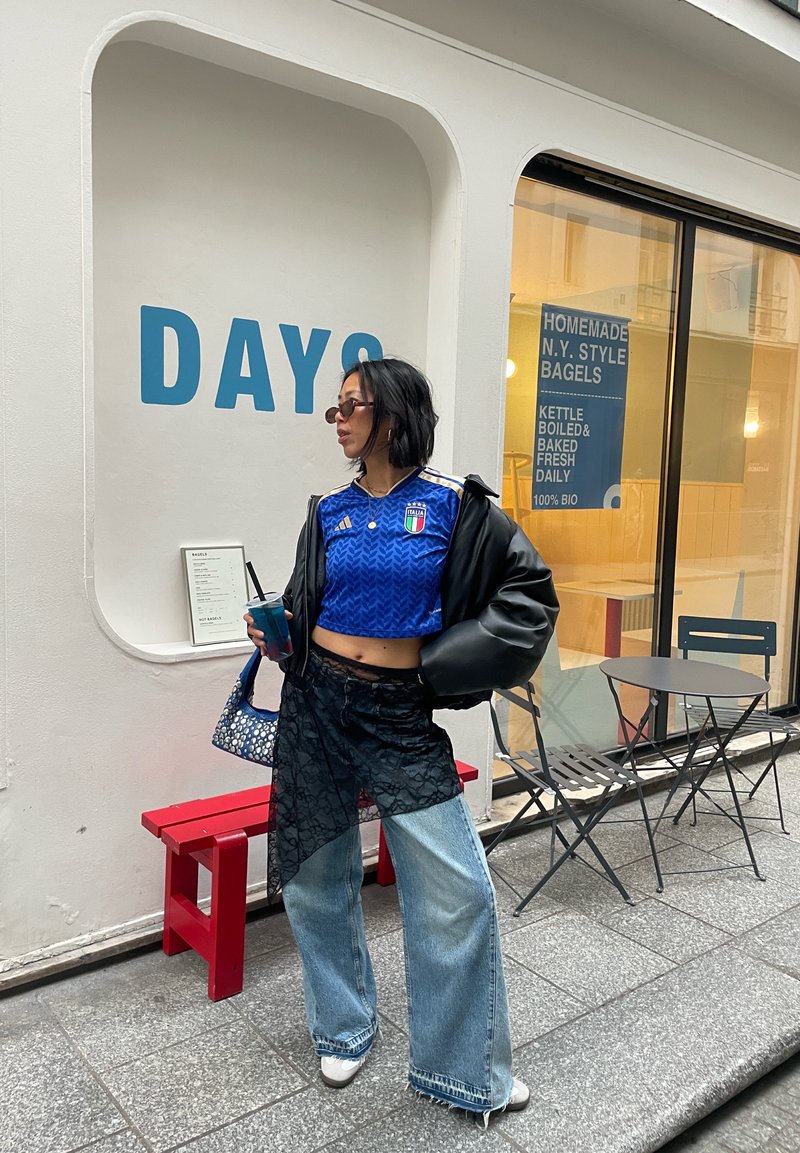 Femme en maillot bleu court et jean tenant un verre, debout près d'un banc rouge à l'extérieur d'un café avec un panneau « DAYS » et un menu de bagels sur le mur.