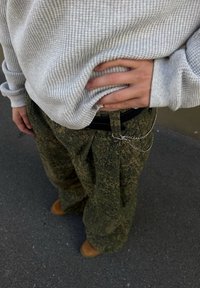 Locker sitzende, olivgrüne gemusterte Hose mit Falten, kombiniert mit einem hellgrauen strukturierten Pullover und einem schwarzen Gürtel mit Kettenakzent.