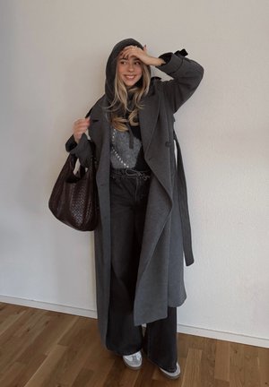 Abrigo de lana gris con capucha y lazos laterales, combinado con un suéter negro y jeans oscuros. Los accesorios incluyen un bolso tejido marrón y zapatillas grises.