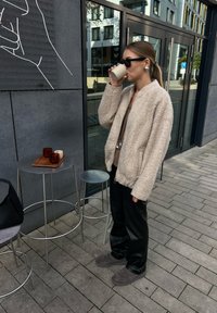 Chaqueta bomber de pelaje suave en beige claro, pantalones de piel sintética negros y botines grises. Sosteniendo una taza beige, de pie cerca de taburetes de metal y una bandeja de madera.
