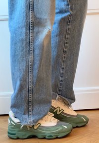 Πράσινα και λευκά αθλητικά παπούτσια New Balance με υφή από δίχτυ και σουέτ, φορεμένα με ανοιχτό μπλε, σκισμένα τζιν παντελόνια με φθαρμένα τελειώματα.