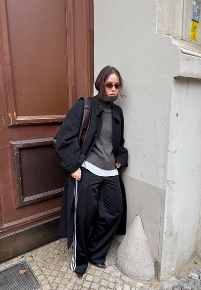Abrigo negro de lana, suéter de tortuga gris con canalé, camisa blanca y pantalones jogger negros con rayas blancas. Bolso de hombro marrón, gafas de sol redondas.