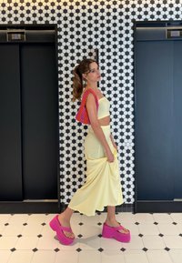 Modell iklädd en åtsittande gul tvådelad outfit med en väska mönstrad i rosa och orange samt klarröda platåsandaler, mot en vägg med svart och vit kakel.