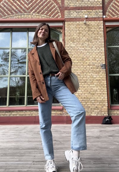 Blazer de pana marrón sobre un suéter verde, jeans rectos azules y zapatillas blancas. El fondo presenta una pared de ladrillo y ventanas verdes.