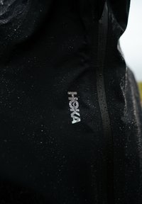 Gros plan sur une veste noire imperméable avec des gouttes de pluie sur le tissu et une fermeture éclair verticale à côté d'un logo HOKA réfléchissant.