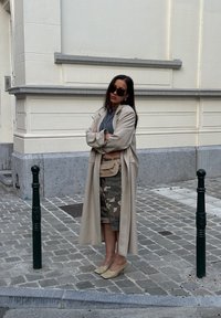Beige lange jas over een grijze top, camouflage shorts en beige hakken. Accessoire: kleine beige pouch riem. Donkere zonnebril, staand op kasseien.
