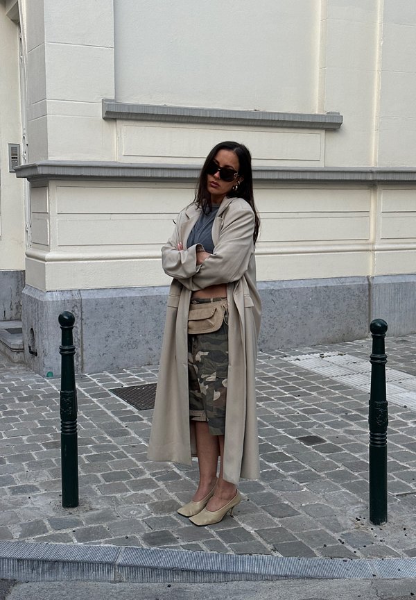 Beige dlouhý kabát přes šedý top, maskáčové šortky a béžové podpatky. Doplňky: malý kožený pásový pytlík. Tmavé sluneční brýle, stojící na dlažebních kostkách.