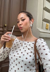 Biała bluzka w brązowe kropki, trzymająca przezroczysty napój z lodem z rurką. Brązowa torba na ramię, białe słuchawki i prosta złota naszyjnik widoczne.