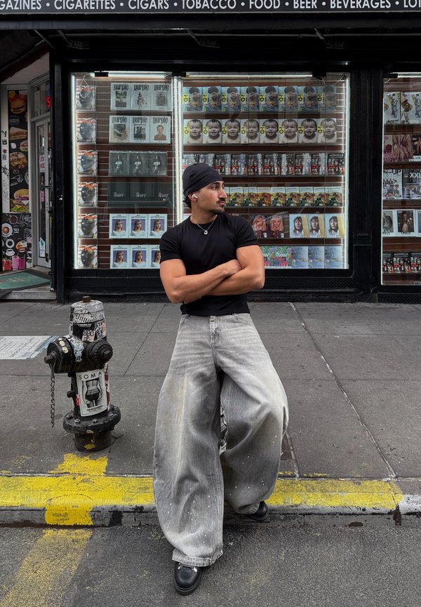 Eine Person trägt ein schwarzes, eng anliegendes T-Shirt und übergroße hellgraue Jeans mit Farbspritzern und steht neben einem strukturierten schwarzen Hydranten.