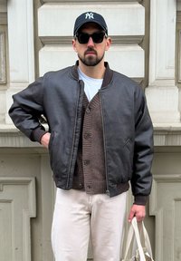 Mees seljas must beisbolimüts, mustad prillid, pruuni nahkjope, pruuni nööbiga sviiter, valged püksid, hoides kreemjat kott.