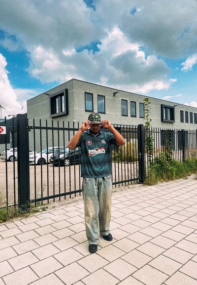Persona que lleva una camiseta de color verde oscuro con un patrón y un logo, junto con unos pantalones ligeros de color gris claro. Lleva una gorra de camuflaje en la cabeza. Pavimento de ladrillos.
