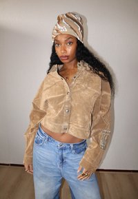 Przycięta brązowa zamszowa kurtka z srebrnymi guzikami i logo Adidas na rękawie, w zestawie z jasnoniebieskimi, luźnymi dżinsami i wzorzystą czapką beanie.