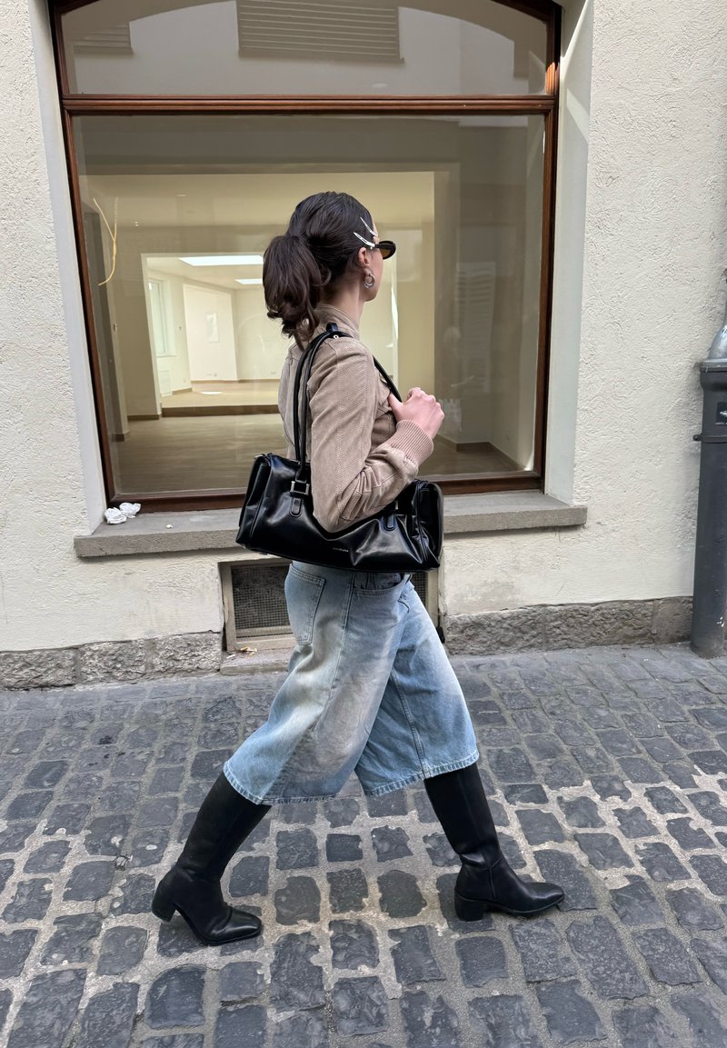 Femme portant des lunettes de soleil, un pull beige, une jupe en jean claire, des bottes noires et un sac à main noir, marchant sur une rue pavée devant une vitrine vide.