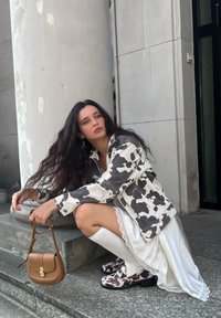 Cow-print jacket, white lace-trimmed dress, thigh-high white boots; brown handbag; black-and-white patterned shoes; natural stone backdrop.