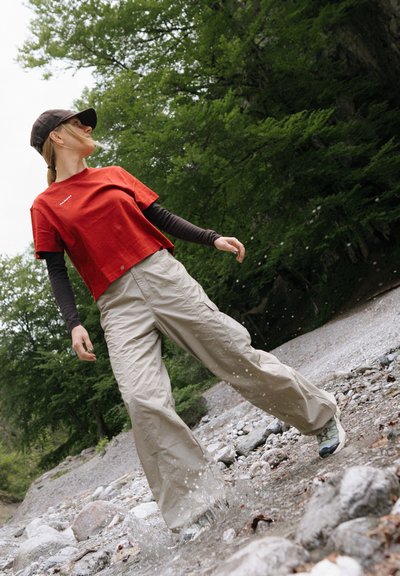 Persona con camisa roja, pantalones beige y gorra caminando por un lecho rocoso de río con agua salpicando, rodeada de árboles verdes.