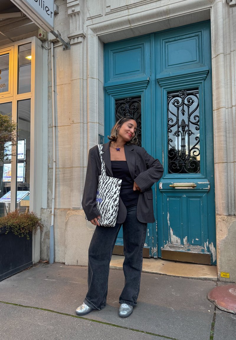 Blazer preto risca de giz, top preto, jeans largos pretos, sapatos prateados. Bolsa com padrão de zebra e detalhes azuis. Portas teal ao fundo.
