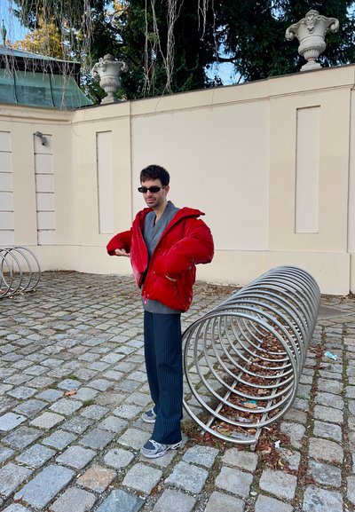 Una chaqueta roja acolchada sobre un suéter gris, combinada con pantalones de rayas oscuros y deportivas grises. Un soporte de bicicleta de metal sobre un suelo de adoquines.