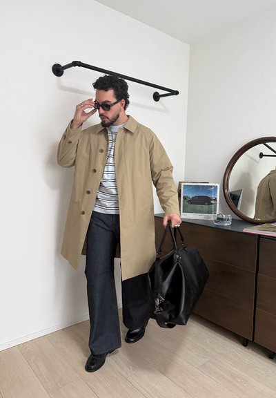 Hombre con abrigo beige, camisa de rayas, pantalones oscuros de pierna ancha y gafas de sol ajustándose las gafas mientras sostiene una gran bolsa negra en el interior cerca de una cómoda con espejo redondo.