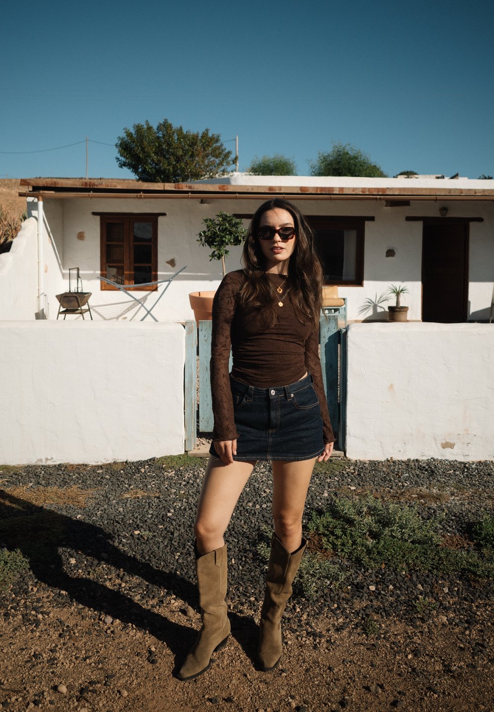 Νεαρή γυναίκα με γυαλιά ηλίου, που φορά καφέ μακρυμάνικη μπλούζα, τζιν φούστα και μπεζ μπότες, στέκεται σε χαλίκι έξω από ένα λευκό σπίτι με γλάστρες.