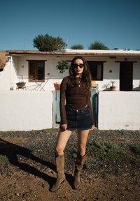 Νεαρή γυναίκα με γυαλιά ηλίου, που φορά καφέ μακρυμάνικη μπλούζα, τζιν φούστα και μπεζ μπότες, στέκεται σε χαλίκι έξω από ένα λευκό σπίτι με γλάστρες.