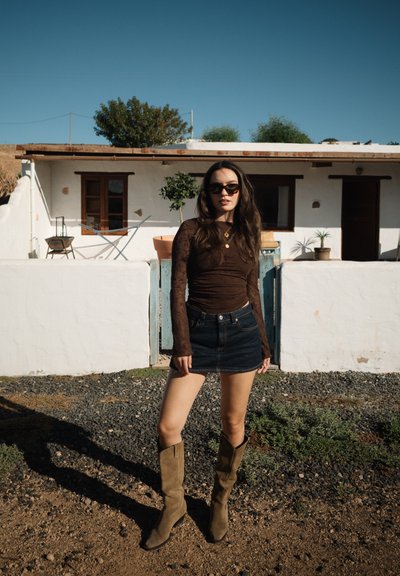 Mujer joven con gafas de sol, vestida con un top marrón de manga larga, falda vaquera y botas beige, de pie sobre grava frente a una casa blanca con plantas en macetas.
