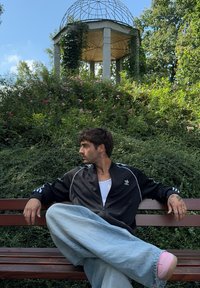 Άνδρας με τατουάζ που φορά μαύρο μπουφάν Adidas και φαρδιά τζιν κάθεται με σταυρωμένα πόδια σε παγκάκι του πάρκου, κοιτώντας στο πλάι, με πράσινο και πέργκολα στο βάθος.